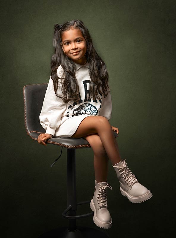 Retrato de niña sentada en una silla de estudio con fondo verde oscuro. Fotografía infantil artística de alta calidad