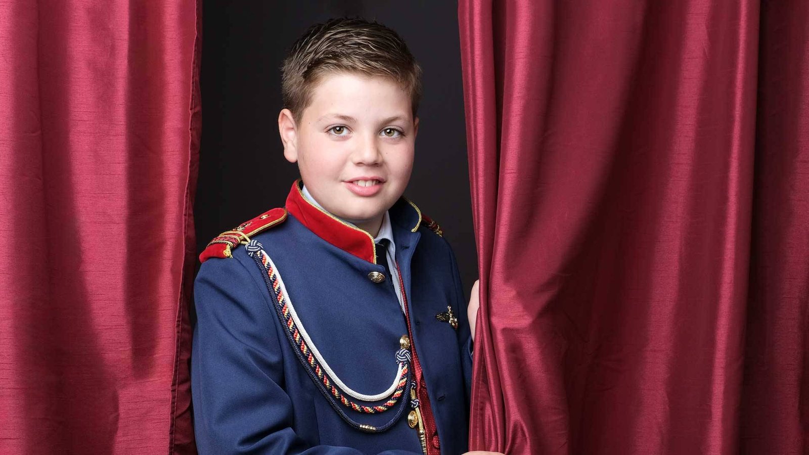 Fotografía de Comunión| Sesiones Únicas y Emotivas Niño de Primera Comunión asomado detrás de una cortina, con traje a juego. Retrato artístico de fotografía de comuniones.
